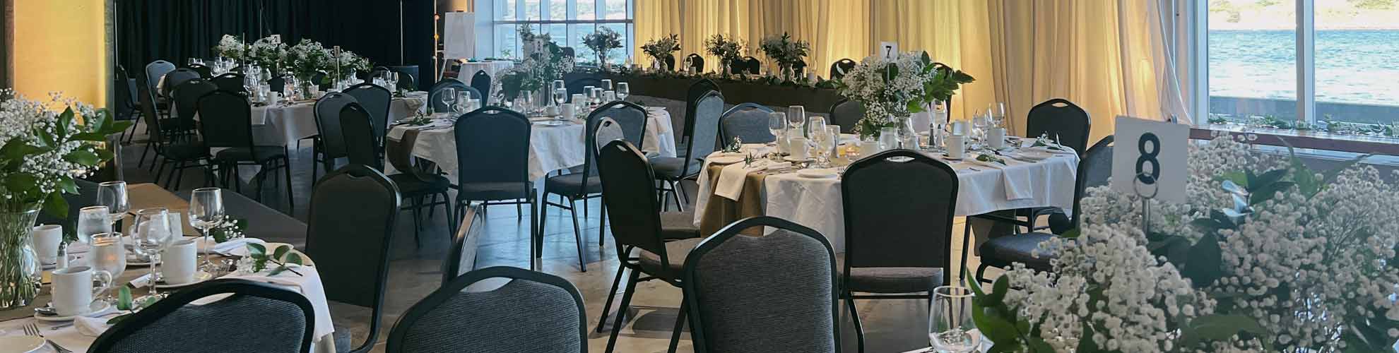 Banquet de mariage avec des tables d’invités ovales décorées et une longue table d’honneur, une petite scène pour un orchestre et de grandes fenêtres allant du sol au plafond. 