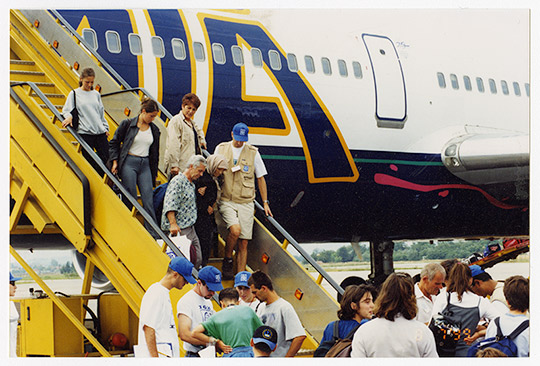 Des réfugiés kosovars à la peau claire débarquent d’un avion en empruntant des escaliers jaunes, tandis que d’autres sont debout à proximité.