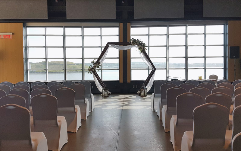 Un ensemble de cérémonie de mariage et des chaises de banquet faisant face à des fenêtres allant du sol au plafond et donnant sur le port et l’île. Une tonnelle hexagonale décorée est placée au centre des fenêtres.