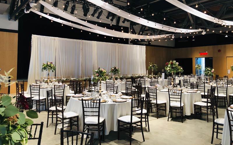 Une salle aménagée en vue d’un mariage et offrant des guirlandes lumineuses et des rideaux blancs au plafond, une toile de fond derrière la table d’honneur et des tables ovales pour les invités.