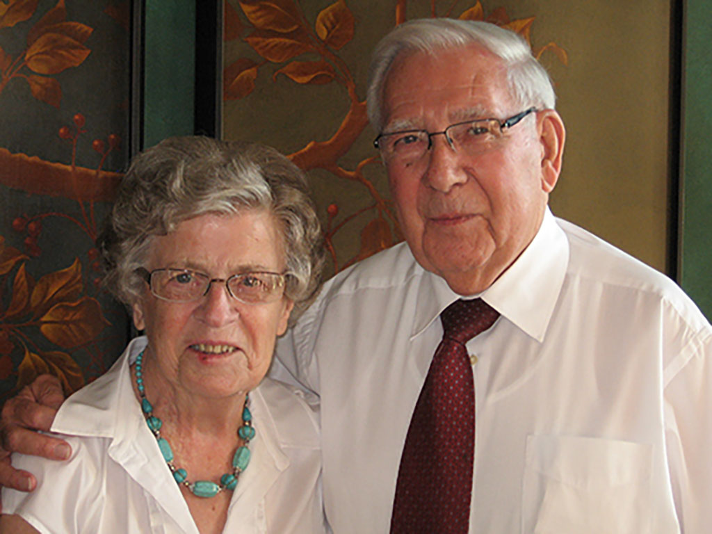 Un portrait en couleur montrant un couple âgé. Ils portent des lunettes et ils se tiennent dans leurs bras.