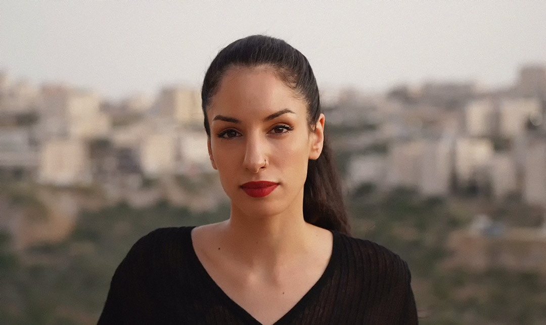 Portrait de Rana Nazzal Hamadeh sur un fond extérieur flou, portant une queue de cheval et un rouge à lèvres rouge.