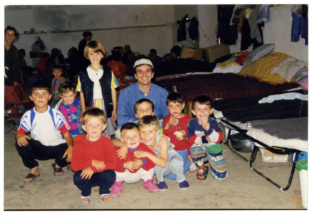Un homme et plusieurs enfants sourient. Ils sont agenouillés à côté d’une rangée de lits de camp.