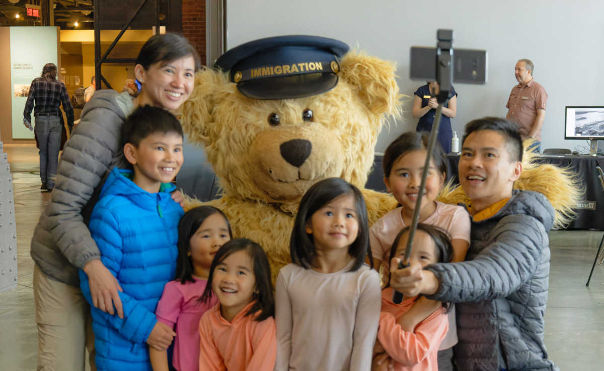 Un grand ours souriant, coiffé d’une casquette portant l’inscription « Immigration », s’agenouille, entouré de six jeunes enfants, d’une femme et d’un homme tenant une perche à égoportrait.