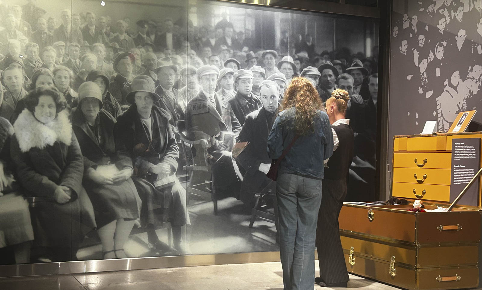 Deux femmes, le dos tourné, devant une image historique occupant tout un mur, représentant une grande foule qui attend sur des bancs.