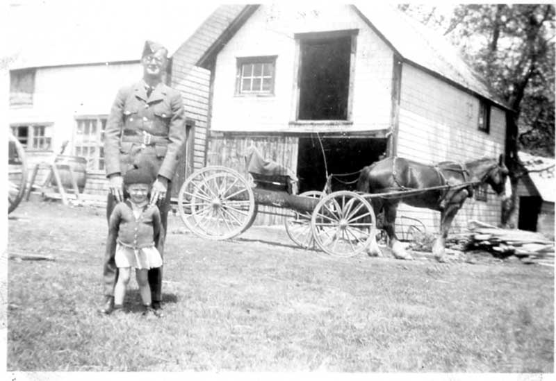 Un homme en uniforme se tenant avec une petite fille devant une maison, un attelage est derrière eux.