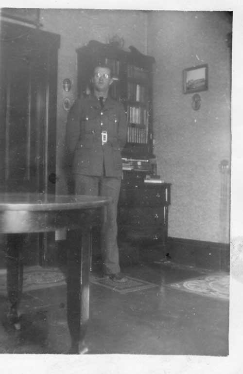 Un homme en uniforme debout dans un coin d’une pièce devant une étagère derrière une table.
