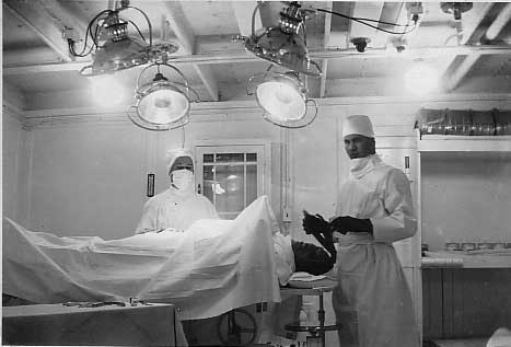 Un homme est allongé sur une table d’opération et deux médecins portant des robes blanches, des casquettes et des masques se tiennent à côté de lui.