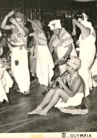 Jeune homme assis au sol, jouant de la flûte alors que des personnes costumées dansent autour de lui.