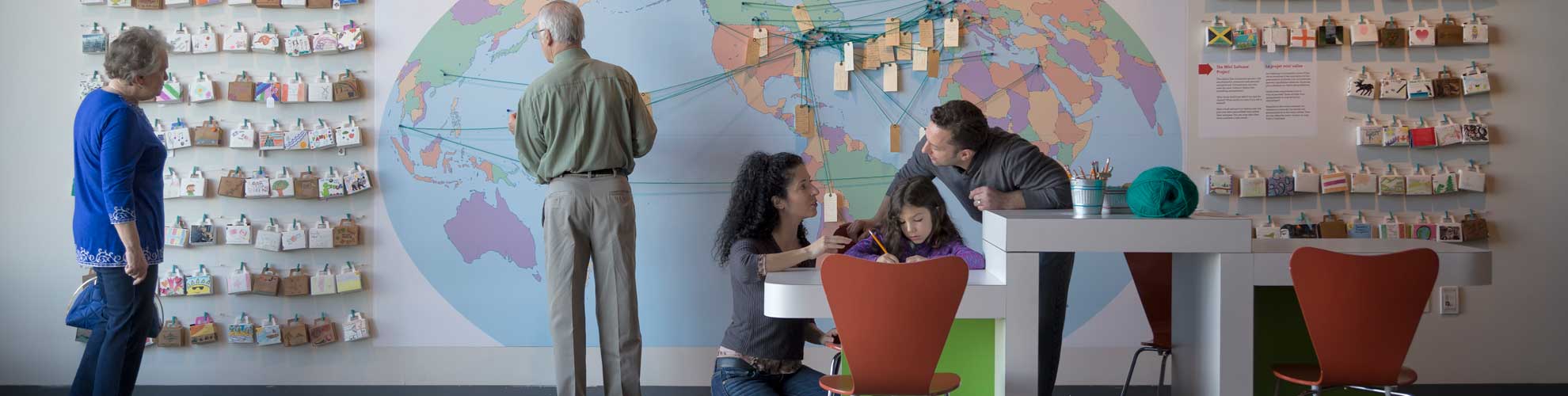 Cinq adultes et un enfant interagissent avec l’exposition, qui présente une carte du monde avec des étiquettes de bagage.