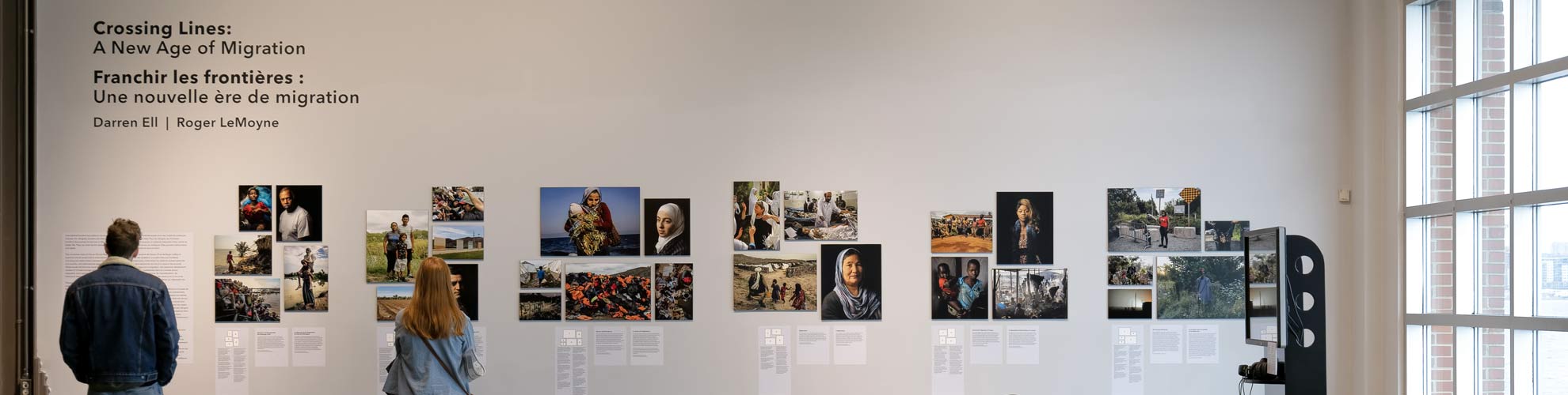 Deux personnes regardent un mur de photographies et de texte dans l’exposition Franchir les frontières.