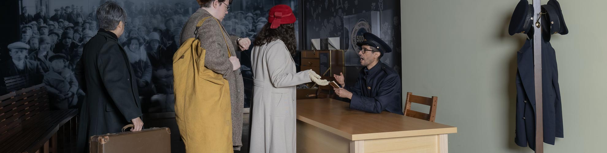 Simulation d’immigration montrant un homme en uniforme à un bureau parlant à trois personnes alignées transportant de vieilles valises.