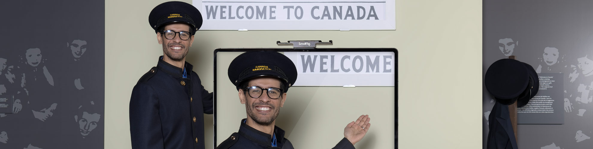 Un homme souriant, en uniforme, au teint moyen, fait un geste vers un panneau « Bienvenue au Canada », debout devant une caméra.