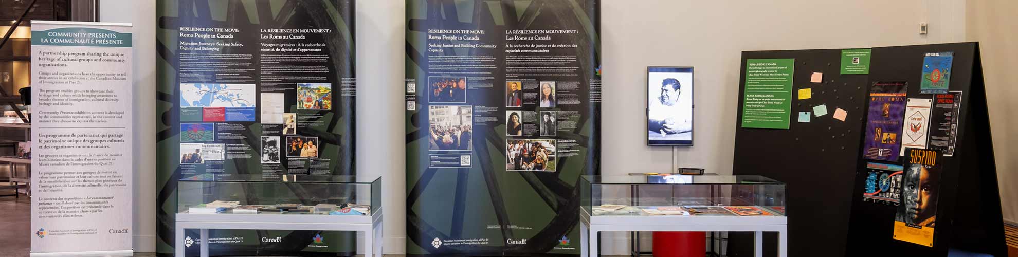 Exposition sur les Roms au Canada avec des panneaux d’information, deux expovitrines et un tableau noir avec des affiches et des notes.