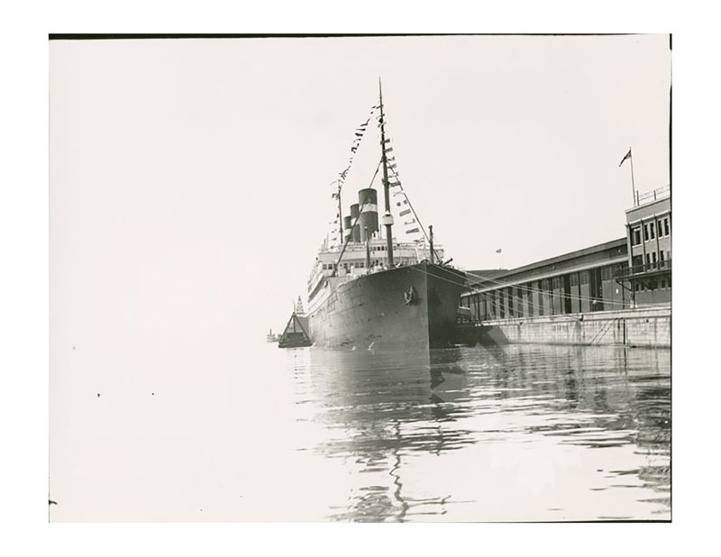 Image en noir et blanc du SS Belgenland amarré à un quai, tel que vu depuis la proue, avec un bâtiment du port visible à droite.