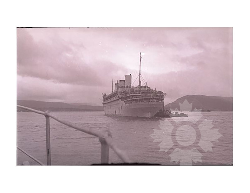 Photo en noir et blanc du SS Île-de-France à quai, vu depuis la proue, avec un remorqueur à ses côtés et des collines côtières en arrière-plan.