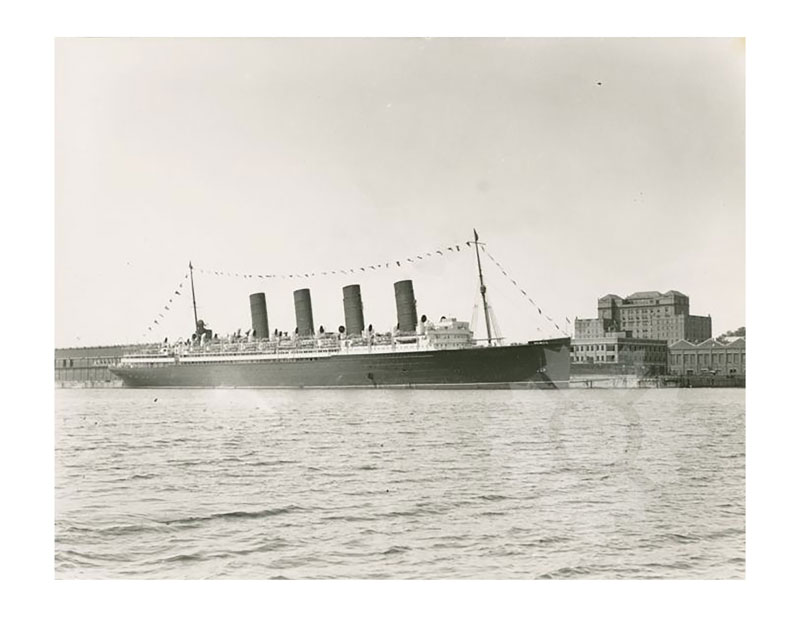 Image en noir et blanc du RMS Mauretania I amarré dans un port, vu de bâbord, avec ses quatre cheminées et des guirlandes festives.