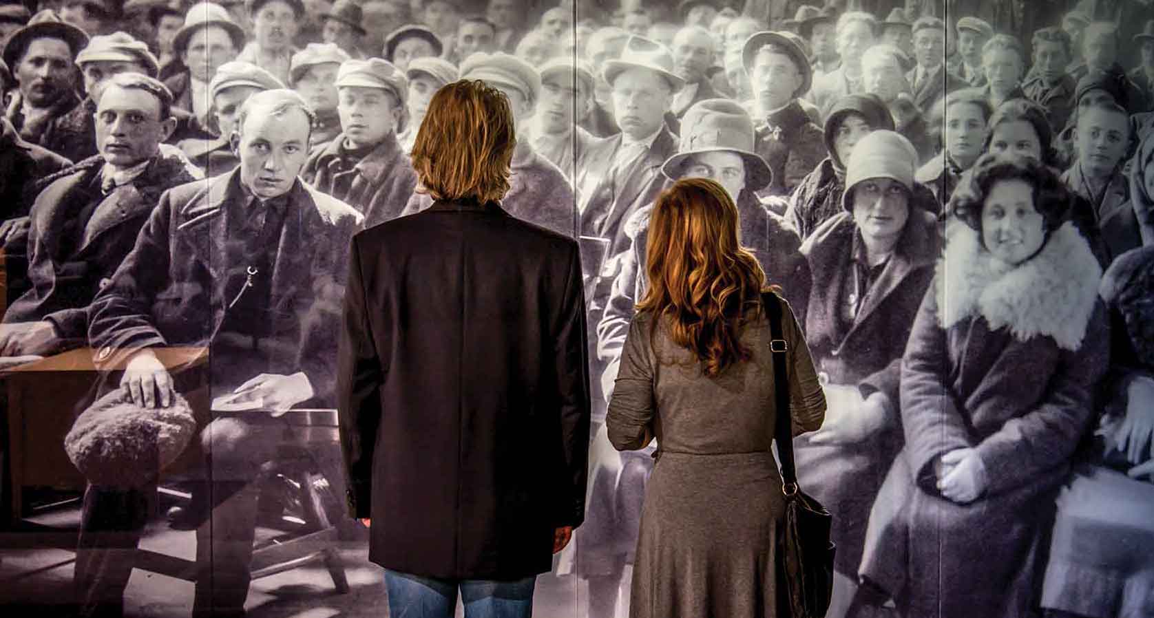 Un homme et une femme regardent une photographie historique grandeur nature d’immigrants qui attendent leur tour.
