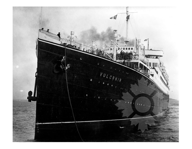 Photo en noir et blanc du MS Vulcania au mouillage, vu de près depuis la proue, avec des passagers visibles sur le pont.