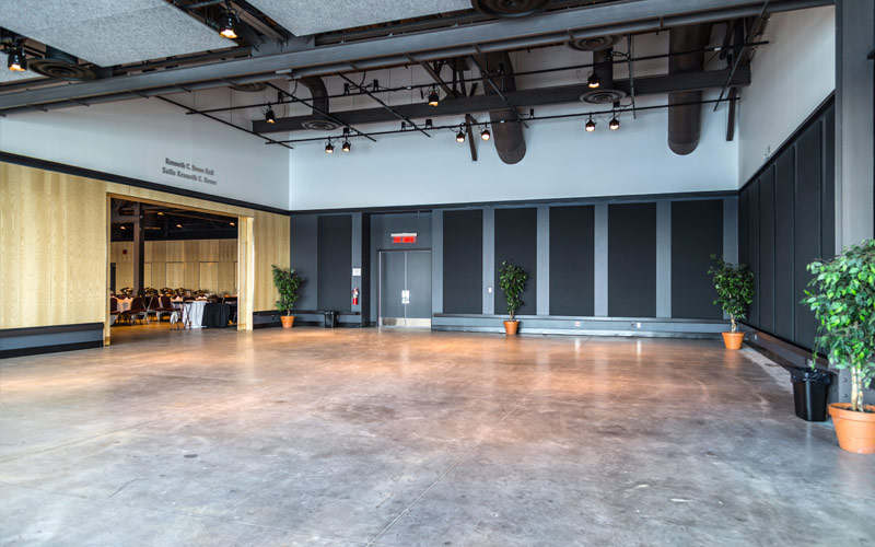 Un grand espace ouvert avec de hauts plafonds et un sol en béton poli. À gauche, l’entrée d’une salle de banquet, et à l’avant et à droite, deux murs équipés de panneaux acoustiques. 
