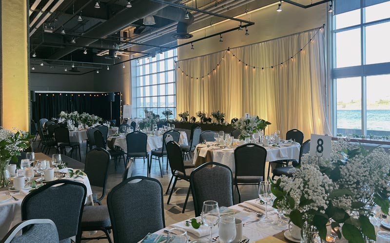 Banquet de mariage avec des tables d’invités ovales décorées et une longue table d’honneur, une petite scène pour un orchestre et de grandes fenêtres allant du sol au plafond.