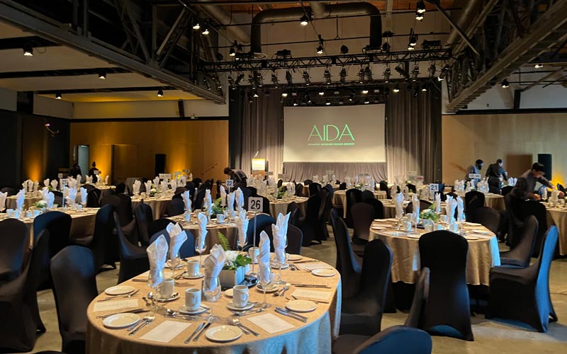 Une salle aux hauts plafonds offre des tables ovales et des chaises de banquet en vue d’un banquet. Sur la scène se trouvent un podium et une toile de projection, les lumières de la scène étant concentrées sur le podium.