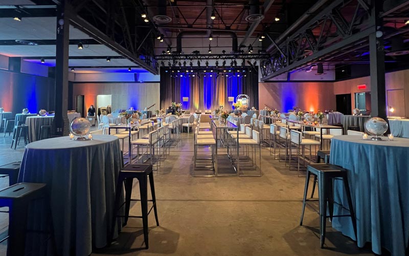 De hautes tables de bar transparentes et des tables à cocktail ornées de tissu bleu offrant des tabourets de bar blancs pour s’asseoir. Les tables sont décorées de nappes bleues, d’un centre de table en forme de globe et de vases-bouteille, et font face à une scène éclairée par des lumières bleues.