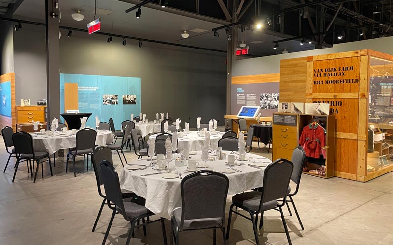 Un banquet situé dans un espace d’exposition du Musée. Des tables ovales sont dressées avec des chaises de banquet, et l’arrière-plan comprend une malle et une valise contenant des vêtements pour enfants.