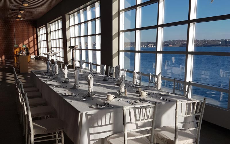 Une longue table et des chaises sont disposées en vue d’un banquet de vingt personnes et plusieurs grandes fenêtres offrent une vue sur le port.