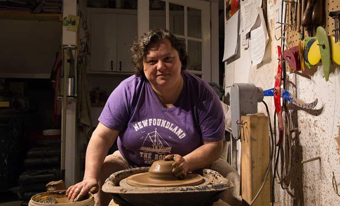 La femme en tee shirt violet est assise dans un atelier, jetant de l’argile céramique sur une roue de poterie.