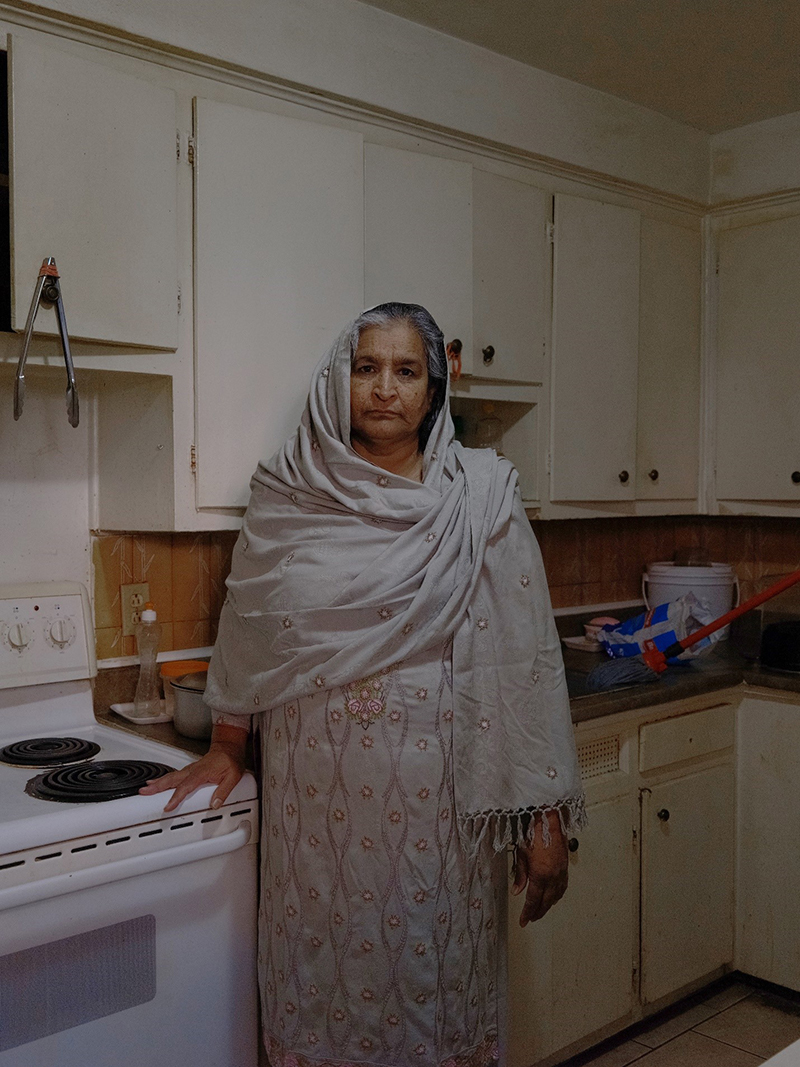 Une femme se tient dans une cuisine, elle porte une robe et un foulard sur la tête et a sa main reposant sur le dessus du poêle.