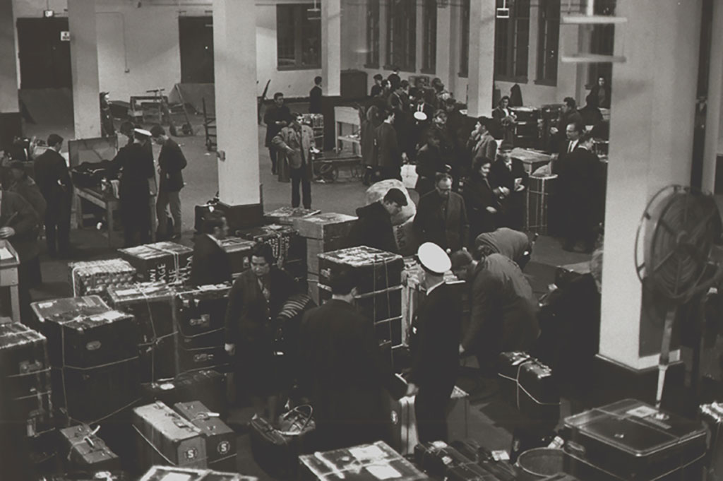 Image archivistique d’une salle d’immigration, avec de nombreux grands troncs, comme vu d’en haut.