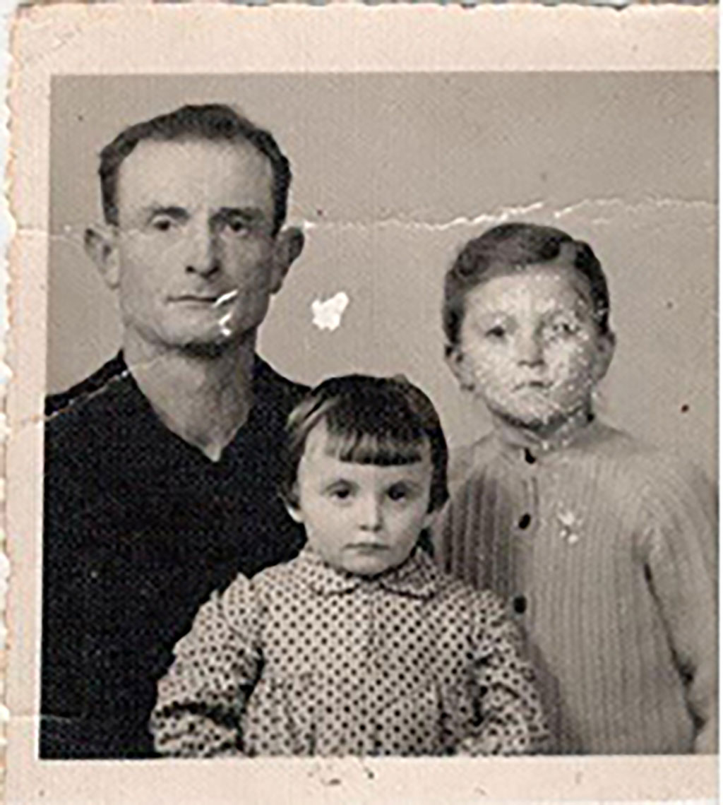 Une vieille photo fissurée montrant un homme portant un chandail noir. Il est assis près d'un petit garçon et d'une petite fille.