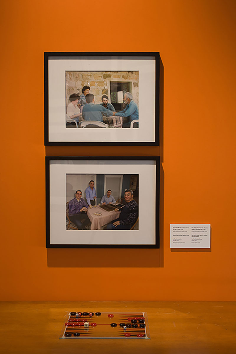 Un mur de couleur orange sur lequel se trouvent deux portraits montrant des gens qui jouent à un jeu de société. Un jeu de société semblable est placé sur le plancher, devant les portraits.