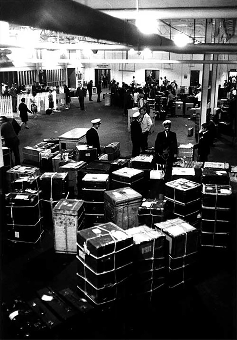Photographie en noir et blanc d'une grande salle remplie de bagages et de douaniers.