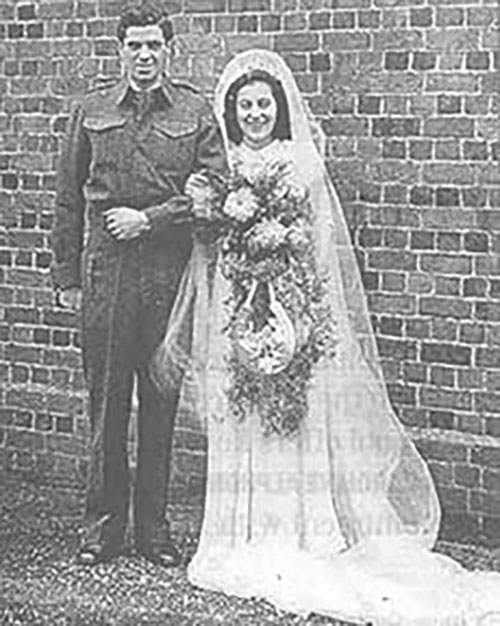 Une image d'archive montrant deux jeunes mariés qui ont les bras enlacés et qui font la pose pour la photo. La mariée tient un magnifique bouquet avec son autre main.