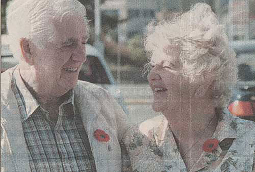 Une photographie en couleur provenant d'un journal. Elle montre un homme et une femme âgés qui portent des coquelicots et qui se sourient l'un l'autre.