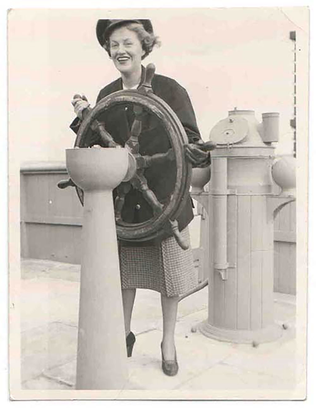 Une femme portant un bonnet de marin tient la barre d'un bateau et affiche un grand sourire.
