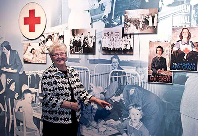 Une femme plus âgée pointe en direction d'une grande murale où se trouve une représentation d'elle-même lorsqu'elle était plus jeune et qu'elle travaillait comme infirmière.