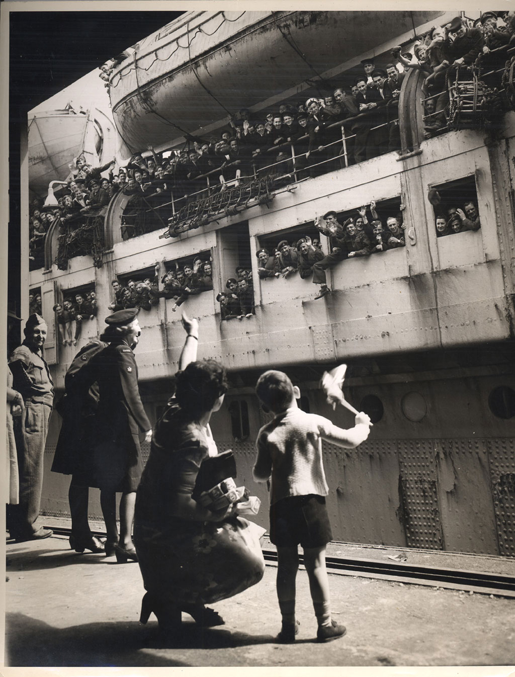 Une vieille photo en noir et blanc du temps de la guerre montrant un navire de transport de troupes amarré. Des centaines de soldats envoient la main et un petit garçon et sa mère leur envoient la main en retour à partir d'une plateforme. La photo montre aussi un militaire qui regarde la scène.