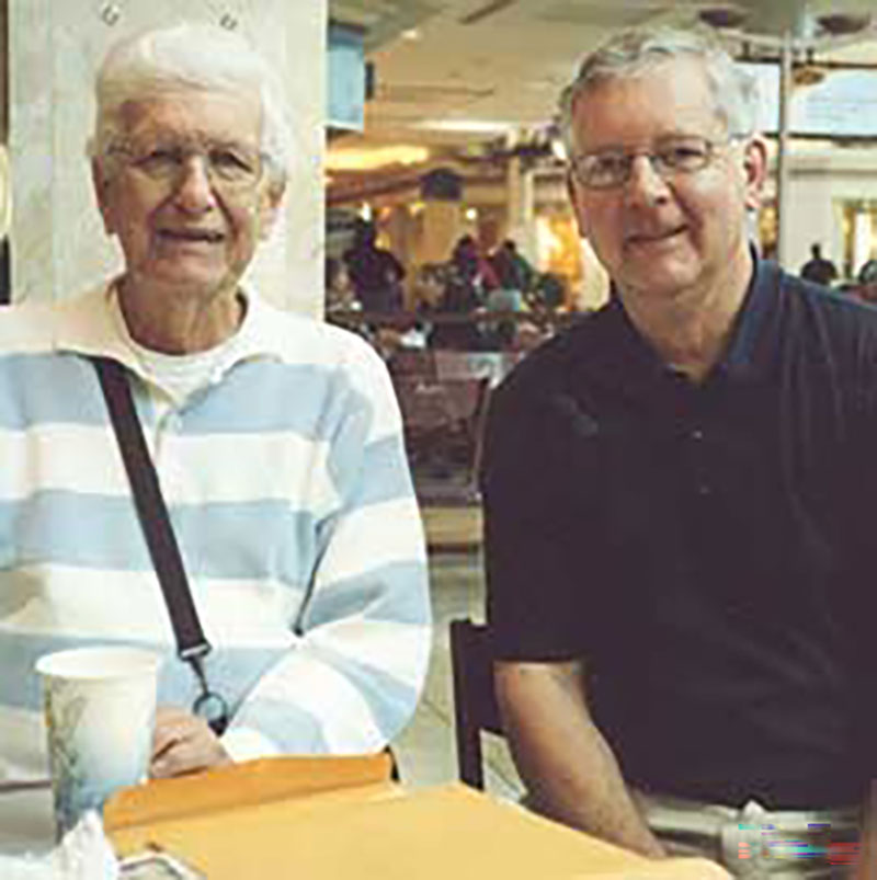 Deux hommes à lunettes sont assis à une table et sourient directement en direction de l'appareil photo.