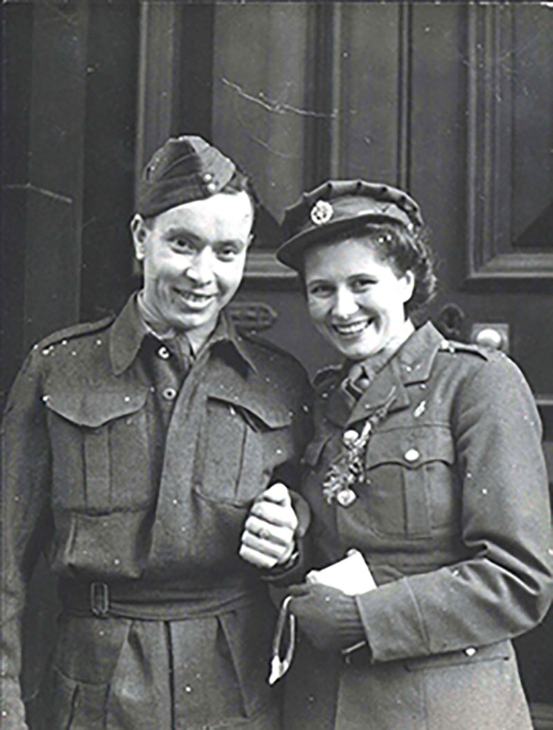 Un jeune homme et une jeune femme qui se tiennent par les mains. Ils portent des chapeaux et des uniformes militaires et sourient en direction de l'appareil photo.