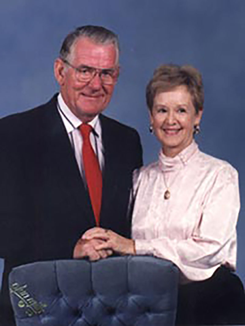 Portrait en couleur d'un homme et d'une femme qui se tiennent debout derrière une chaise bleue. Ils se tiennent la main sur la chaise. L'homme porte un costume et une cravate rouge, la femme porte une blouse d'un rose tendre et l'arrière-plan est bleu.