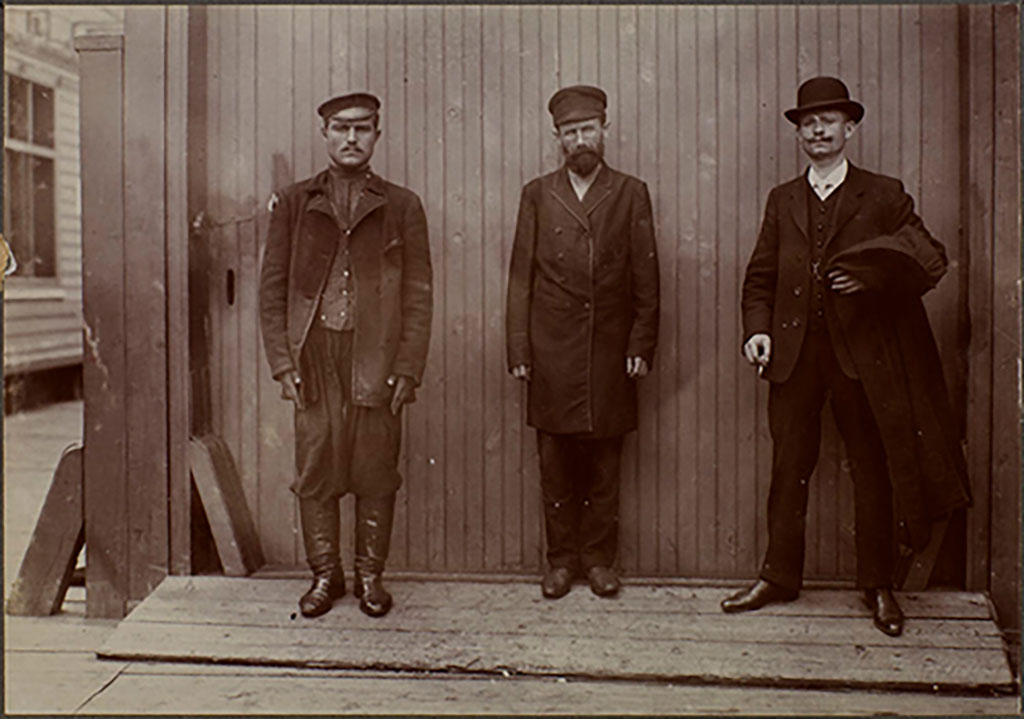 Image d'archive montrant trois hommes debout devant un mur. Ils portent tous des vêtements d'époque.