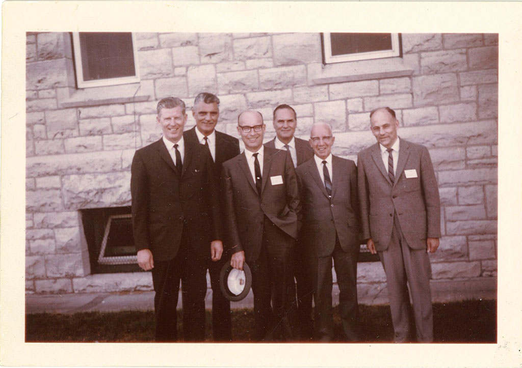 Un groupe d’hommes vêtus de costumes et de cravates se tient devant un bâtiment en pierre tandis qu’ils sourient pour la caméra.