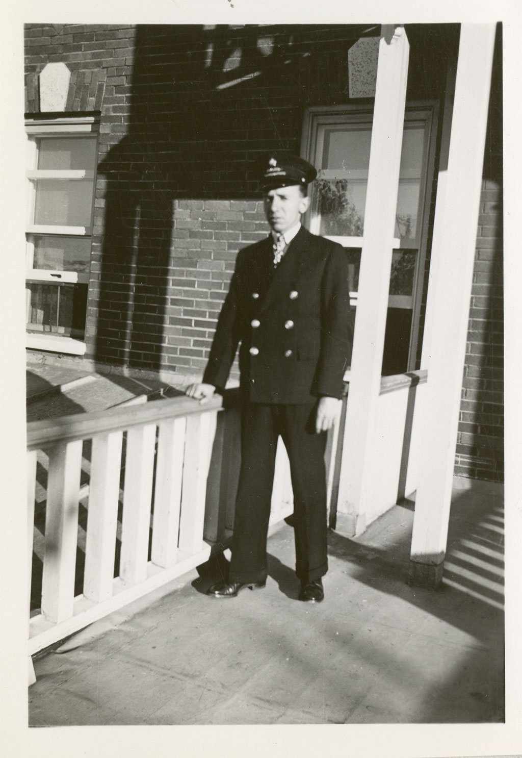 Un homme en casquette et en uniforme se tient la main sur une rambarde en bois en regardant la caméra.