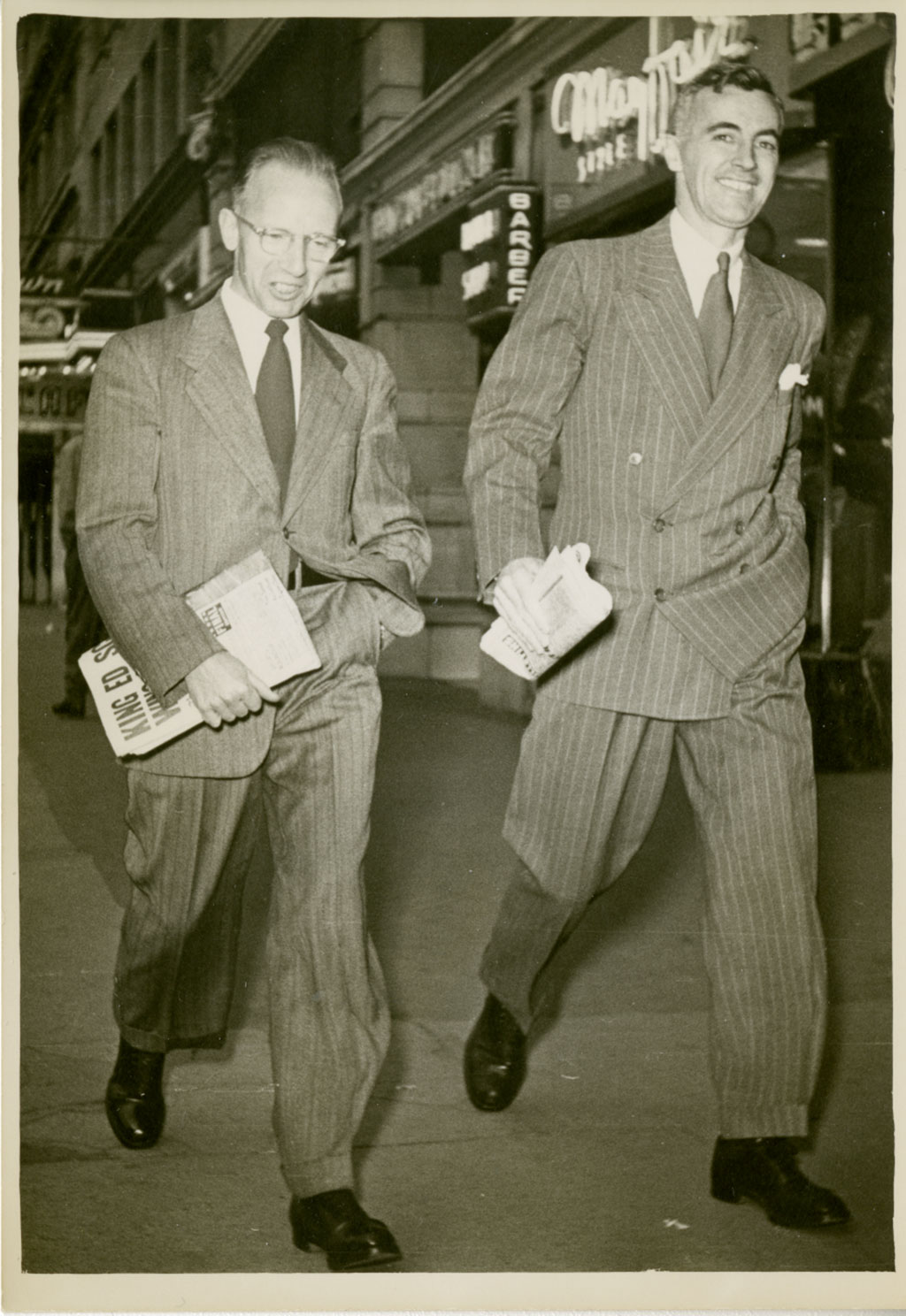 Deux hommes marchent avec détermination, tenant les journaux dans une main et l’autre main dans leurs poches.