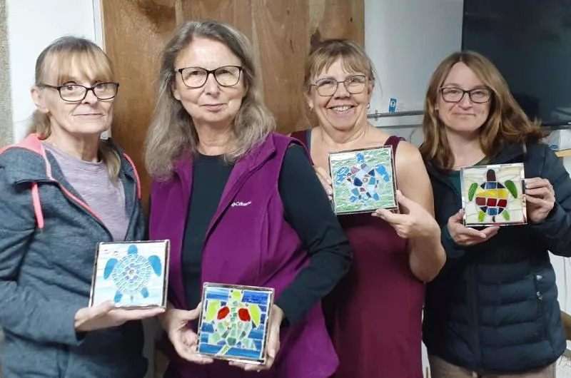Quatre femmes participant à un atelier d’art du verre présentent leurs panneaux de mosaïque de tortue de mer en vitrail faits à la main.