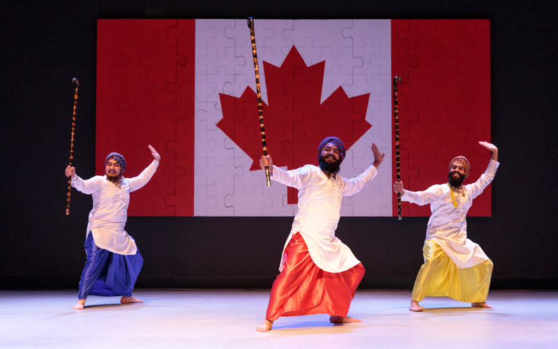 Trois hommes dansent devant un grand drapeau canadien avec un motif de pièces de casse-tête.