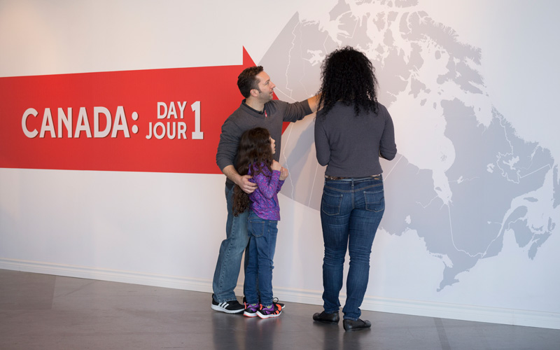 Deux adultes et un enfant regardent une grande carte du Canada sur le mur. Un panneau rouge portant le titre de l’exposition se trouve derrière eux.